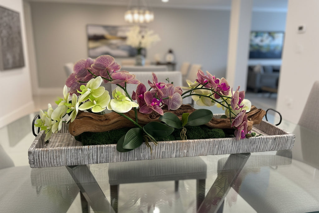 A long rectangular woven tray centerpiece filled with artificial orchids in white and pink-purple tones, green leaves, and a piece of driftwood, displayed on a table in a dining room setting.